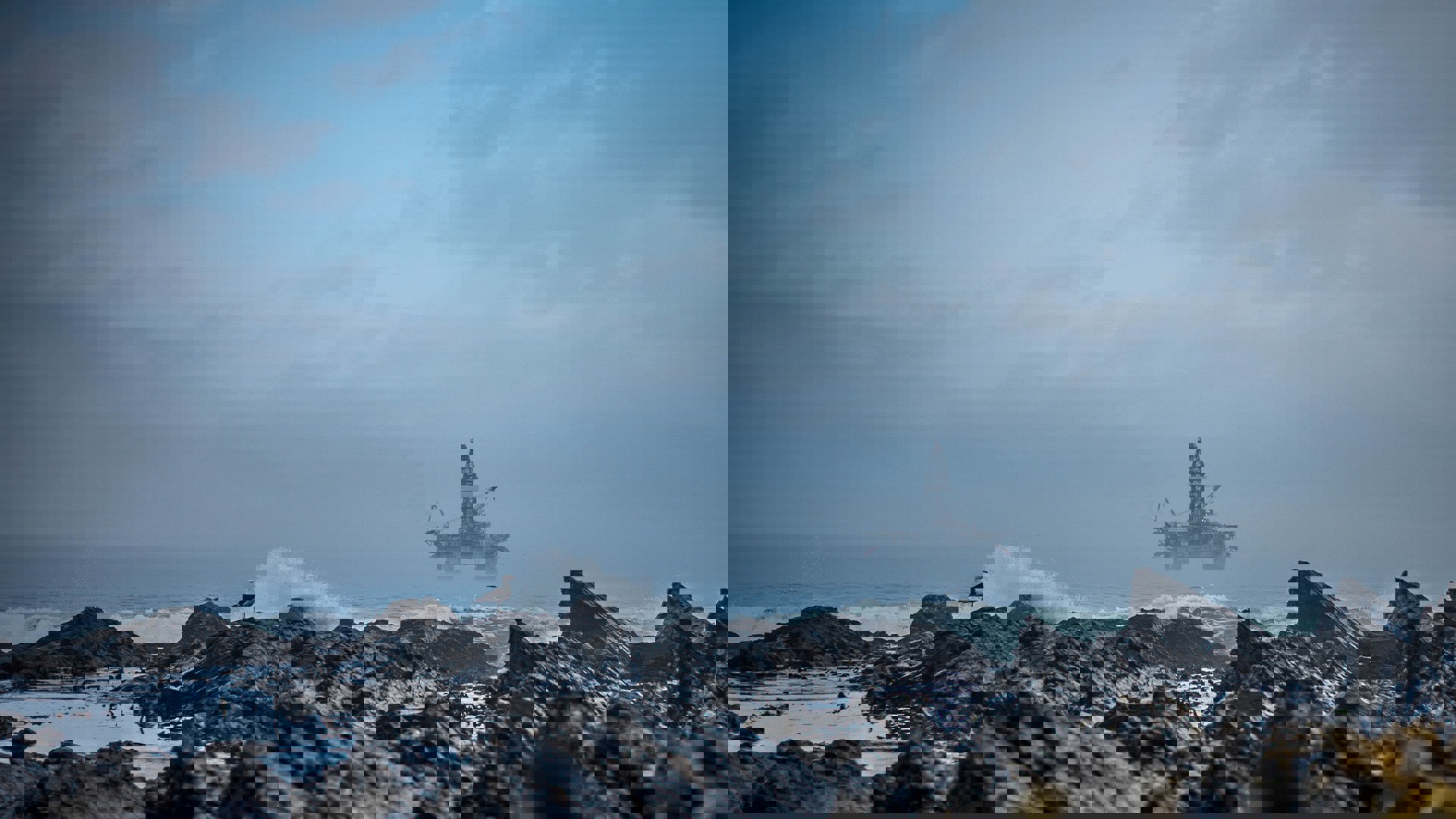 An oil rig stands in the ocean, surrounded by rocky formations and waves, illustrating industrial activity at sea.